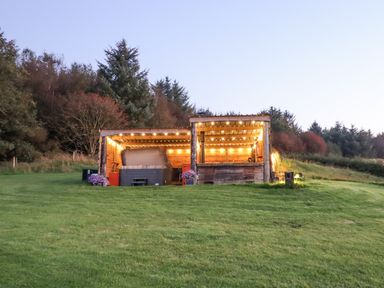 A wooden outdoor structure with string lights on a grassy hill surrounded by trees at Rhosydd in Cyffylliog near Ruthin