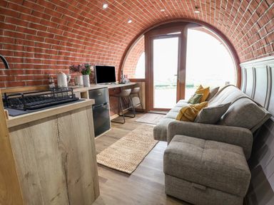 A curved brick room with a grey sofa and ottoman opposite a counter with a monitor and bar stools at Cae Bach in Cyffylliog near Ruthin