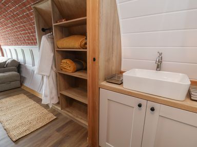 A living space with wooden shelves holding folded yellow blankets white robe hanging a sink with cabinet below and a gray sofa near a brick wall at Cae Bach in Cyffylliog near Ruthin
