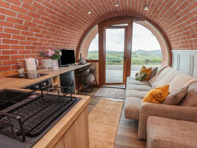 An interior room with a curved brick ceiling a wooden countertop with kitchen appliances a sofa with cushions and a large arched window and door showing a countryside view at Cae Bach in Cyffylliog near Ruthin