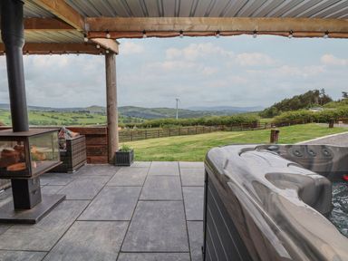 An outdoor covered patio with a fireplace a hot tub seating area and a view of fields and hills at Cae Bach in Cyffylliog near Ruthin