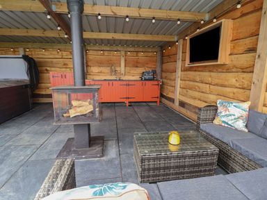 An outdoor covered seating area with wicker furniture a glass coffee table a wood burning stove orange cabinets with a sink and grill mounted television on a wooden wall at Cae Bach in Cyffylliog near Ruthin