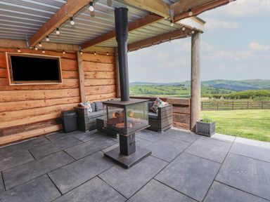 An outdoor covered patio with a wood stove a sofa a wall-mounted tv and string lights overlooking green hills at Cae Bach in Cyffylliog near Ruthin