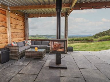 An outdoor covered seating area with a sectional sofa table wood stove and countryside views at Cae Bach in Cyffylliog near Ruthin