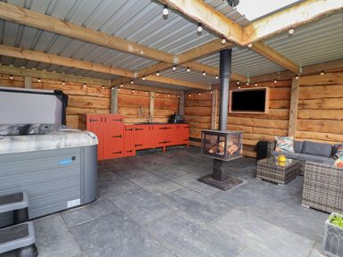 An outdoor covered patio with a hot tub red kitchen cabinets a wood burning stove and wicker seating at Cae Bach in Cyffylliog near Ruthin