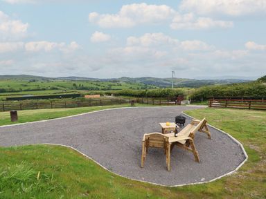 A gravel seating area with wooden chairs a table and a bottle on the table in a grassy outdoor setting with hills and wind turbines in the background at Cae Bach in Cyffylliog near Ruthin