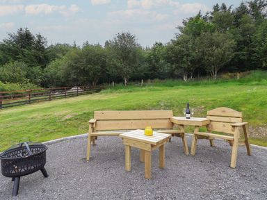 Outdoor seating area with wooden benches a table and a fire pit on gravel surrounded by grass and trees at Cae Bach in Cyffylliog near Ruthin
