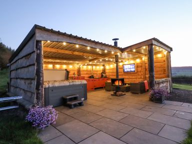 An outdoor covered patio with a hot tub chairs a wood stove string lights and a TV at Cae Bach in Cyffylliog near Ruthin