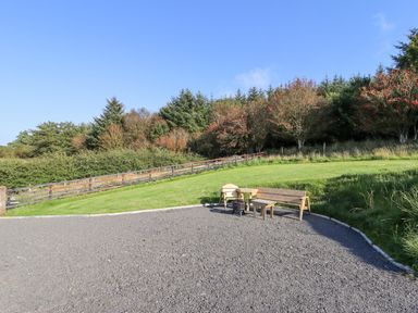 An outdoor area with two wooden benches a small table and a fire pit near a grassy slope and trees at Cae Bach in Cyffylliog near Ruthin