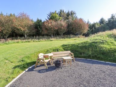 An outdoor seating area with wooden chairs a bench tables and a fire pit surrounded by grass and trees at Cae Bach in Cyffylliog near Ruthin