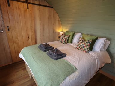 A bedroom with a double bed green and patterned pillows folded towels wooden wall and lamp at Weirglodd in Ruthin
