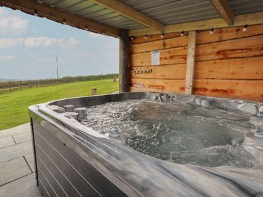 A hot tub on a tiled patio under a wooden and metal roof with a grassy field and wind turbine in the background at Weirglodd in Ruthin