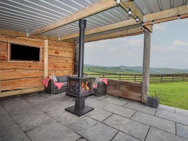 An outdoor covered patio with a wood burning stove seating area and a mounted television overlooking a green field at Weirglodd in Ruthin