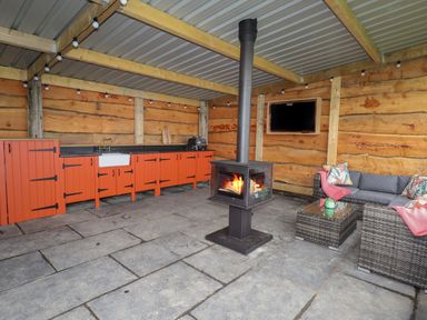 An outdoor kitchen with orange cabinets a wood burning stove a gray wicker sofa and a wall mounted television at Weirglodd in Ruthin