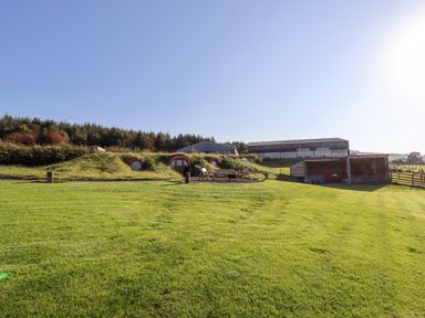 A grassy outdoor area with a hobbit-style house, wooden chairs around a fire pit and farm buildings in the background at Weirglodd in Ruthin