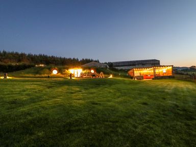 A grassy outdoor area with illuminated round doors in a hillside and a lit wooden shelter at Weirglodd in Ruthin