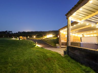 An outdoor area with a covered hot tub lit by string lights and a path leading to benches at Weirglodd in Ruthin