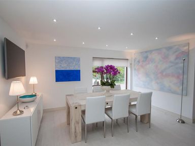 A dining room with a light wood table and white chairs purple flowers on the table paintings on the walls and lamps on a white cabinet at The Arthouse in Ambleside