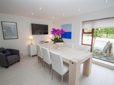 A dining room with a light wooden table white chairs a large flower centerpiece a grey armchair and a wall-mounted television at The Arthouse in Ambleside
