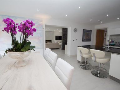 A kitchen with a light wood dining table and purple flowers a kitchen island with two white bar stools and a living room in the background at The Arthouse in Ambleside