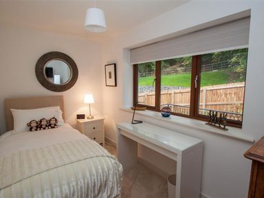 A bedroom with a single bed and three stuffed monkeys on it a nightstand with a lamp and alarm clock a desk below a window with a view of a garden at The Arthouse in Ambleside