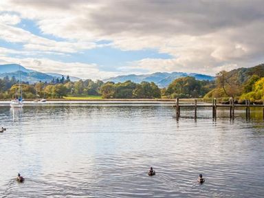 A lake with ducks swimming near a wooden pier and sailboats with hills and trees in the background at The Arthouse in Ambleside