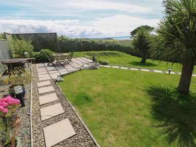 A garden with a paved patio area with table and chairs a grassy lawn palm trees and a view of countryside and water at Carreg Ddu in Solva