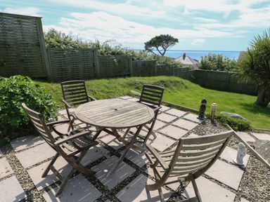 An outdoor patio with a round wooden table and four chairs overlooking a grassy garden with fencing and a distant view of the sea at Carreg Ddu in Solva