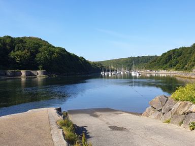 A concrete boat ramp leading into a calm river with sailboats moored in the distance and green hills on either side at Carreg Ddu in Solva