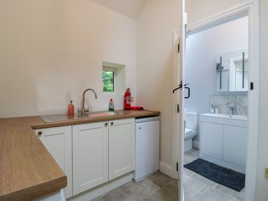 A kitchen area with a sink soap dispenser and cleaning products next to a small fridge and an open door leading to a bathroom at West View in Feltwell