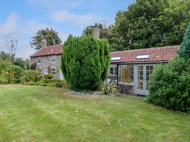 A garden with grass lawn and bushes in front of a stone house with red tiled roof at West View in Feltwell