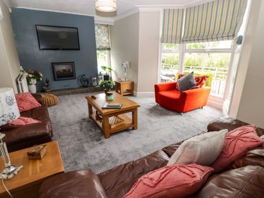 A living room with brown leather sofas a red armchair wooden tables a TV mounted on a blue wall and windows with striped blinds at Morva in New Quay