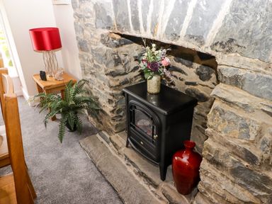 A black stove with a vase of flowers on top set into a stone fireplace next to a red vase and a wooden side table with a red lamp and a potted plant on a carpeted floor at Morva in New Quay
