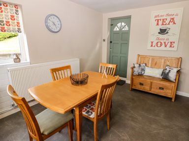 A dining area with a wooden table and four chairs a wooden bench with cushions a green door a wall clock and a window with patterned blinds at Morva in New Quay