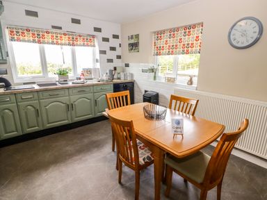 A kitchen with a wooden dining table and chairs next to green cabinets and two windows with patterned blinds at Morva in New Quay