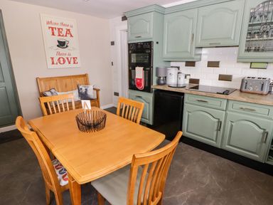 A kitchen with green cabinets a wooden dining table and chairs a wooden bench with cushions and a sign on the wall reading where there is tea there is love at Morva in New Quay