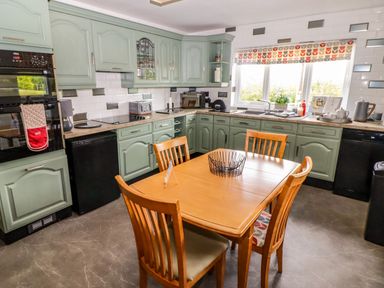 A kitchen with green cabinets wooden dining table and chairs and a window with patterned blind at Morva in New Quay