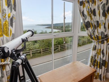A window with a telescope and patterned curtains overlooking a coastal road and sea at Morva in New Quay