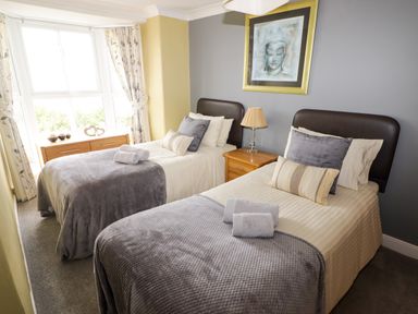 A bedroom with two single beds side by side with grey blankets and pillows a bedside table with a lamp a dresser next to a large window and a framed artwork on the wall at Morva in New Quay