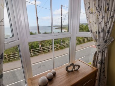 A window with floral curtains overlooking a road and coastline with decorative items on a wooden chest at Morva in New Quay
