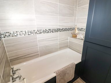 A bathroom with a bathtub tiled walls a towel on the bathtub and a dark cabinet with a sailboat and a jar of shells on top at Morva in New Quay