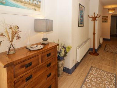 A hallway with a wooden chest of drawers holding a lamp and decorative items a coat rack radiator and floral rugs at Morva in New Quay