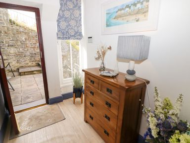 An entryway with a wooden chest of drawers a lamp flowers a window with a patterned blind and an open door showing an outdoor stone patio at Morva in New Quay