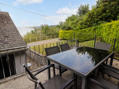 An outdoor patio area with a glass table and six chairs overlooking a road and greenery at Morva in New Quay