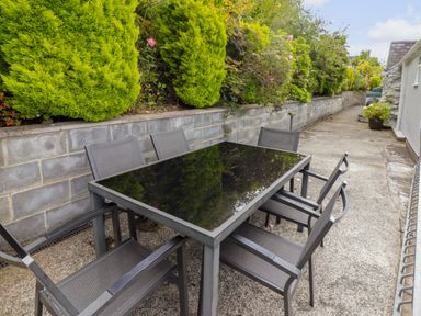 An outdoor patio area with a black glass table and six metal chairs next to a concrete wall with green bushes at Morva in New Quay