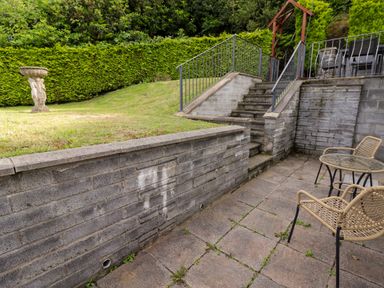 An outdoor patio area with two wicker chairs and a table stone steps with metal railings leading to a grassy area with a bird bath and green hedge Morva New Quay