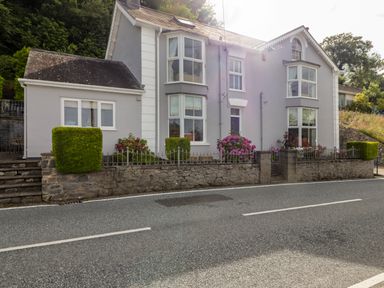 A grey two-story house with bay windows and a stone fence along the road at Morva in New Quay