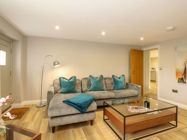 A living room with a gray sectional sofa with teal pillows and blanket a metal floor lamp and a glass coffee table at Daisys Place in South Creake near Little Walsingham