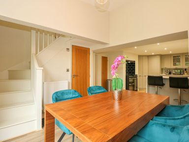 A dining area with a wooden table, blue chairs, a flower vase, staircase, kitchen with bar stools and appliances at Daisys Place in South Creake near Little Walsingham