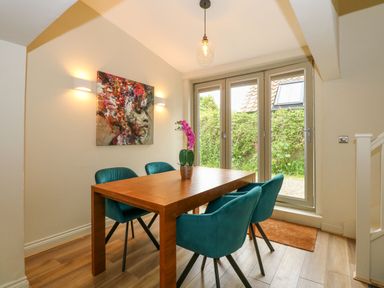 A dining area with a wooden table and four teal chairs near glass doors and a painting on the wall at Daisys Place South Creake near Little Walsingham